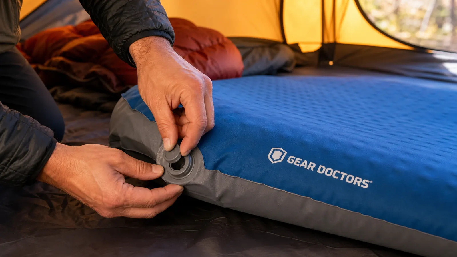 Hands opening the valve on the Gear Doctors Oxylus sleeping pad while it self-inflates on the ground inside a tent